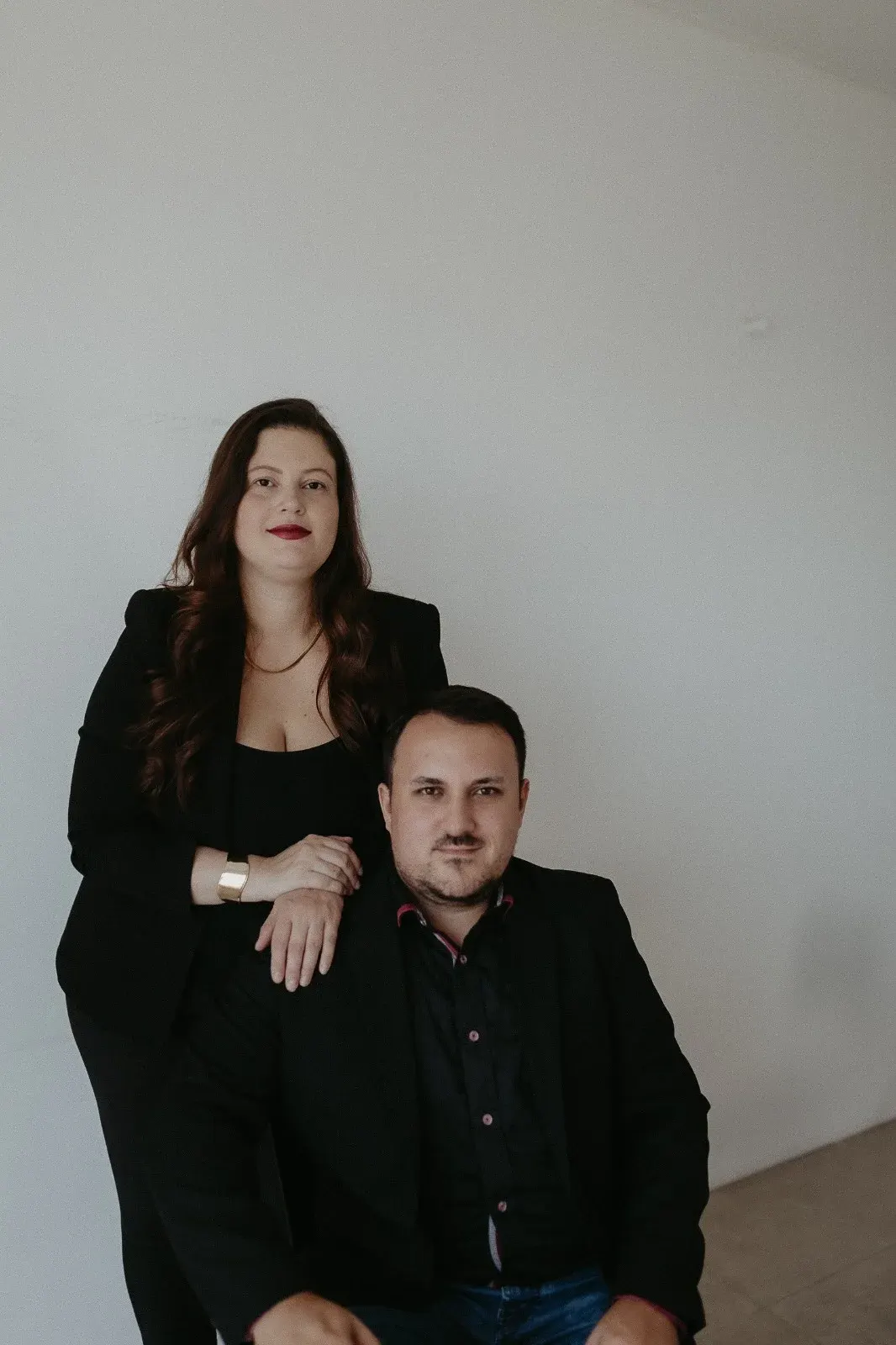 Duas pessoas do escritório, uma em pé e outra sentada, de roupa preta, diante de uma parede clara